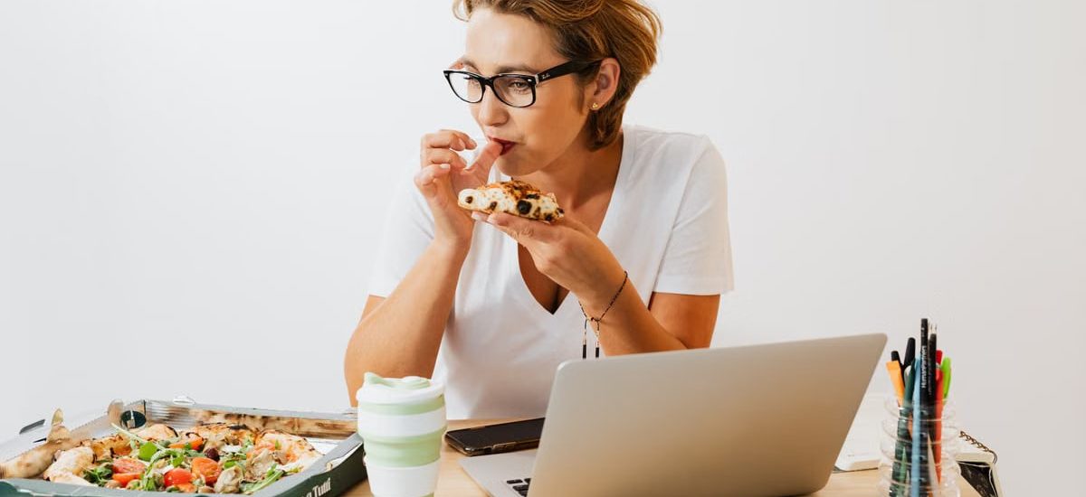 A woman eating pizza at her workspace with a laptop and stationary around.