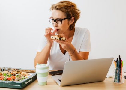 A woman eating pizza at her workspace with a laptop and stationary around.