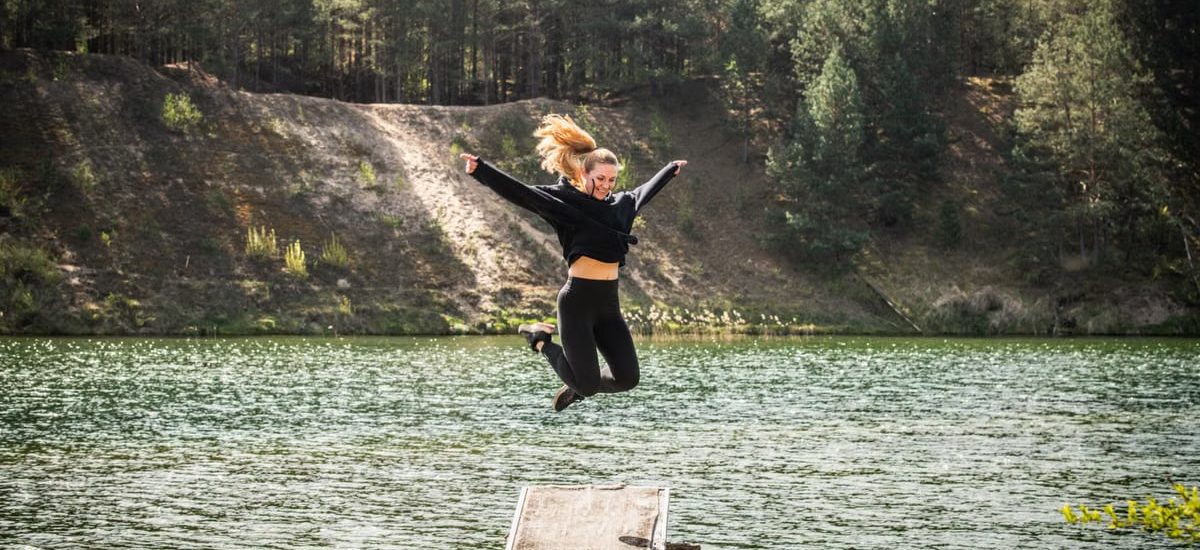 A woman leaps joyfully from a pier into a scenic forested lake in Lithuania, embracing adventure.
