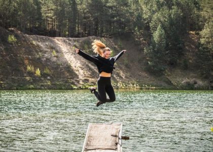 A woman leaps joyfully from a pier into a scenic forested lake in Lithuania, embracing adventure.